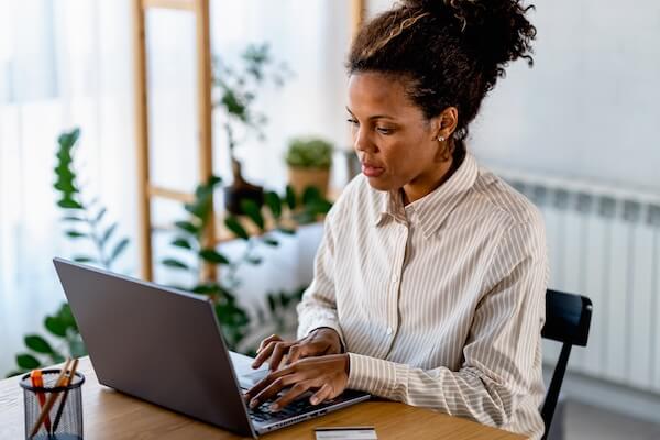 A women wearing a tan and white striped button up typing on a laptop while sitting at a wooden table