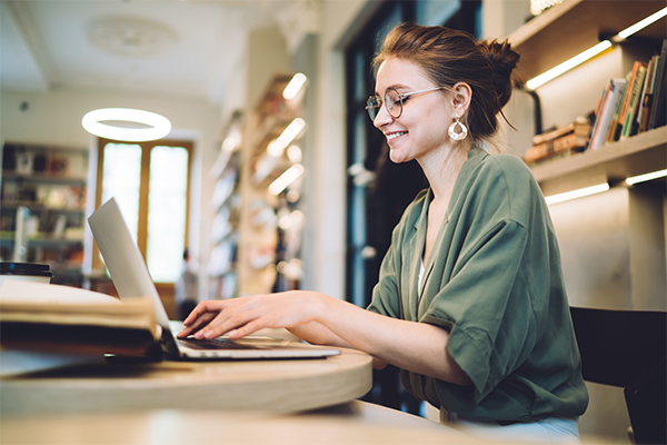 A Young adult woman with brown hair in a low bun wearing a green cardigan, glasses and gold earrings sitting at a table typing on a silver laptop.