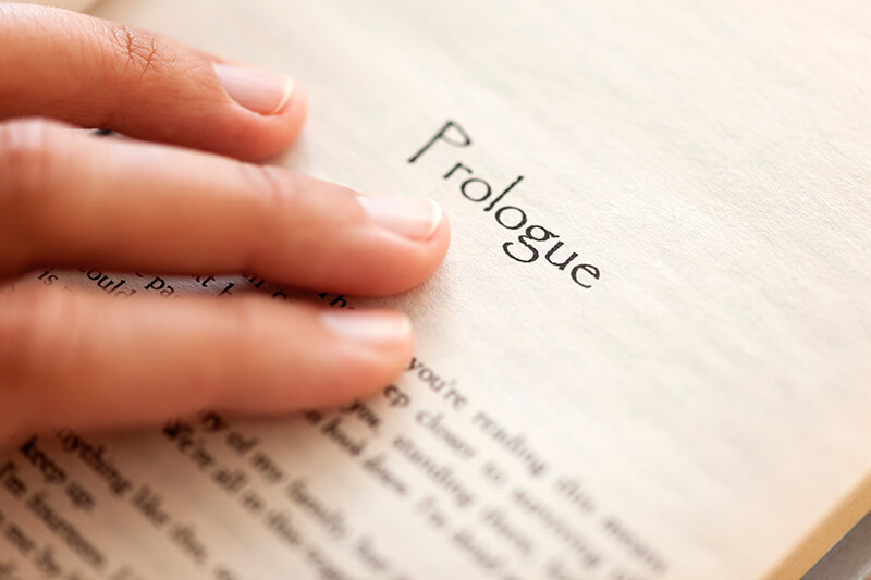 Prologues-vs-Epilogues Close up image of a book page with the text "prologue" at the top. There is a hand touching the page right below the font.
