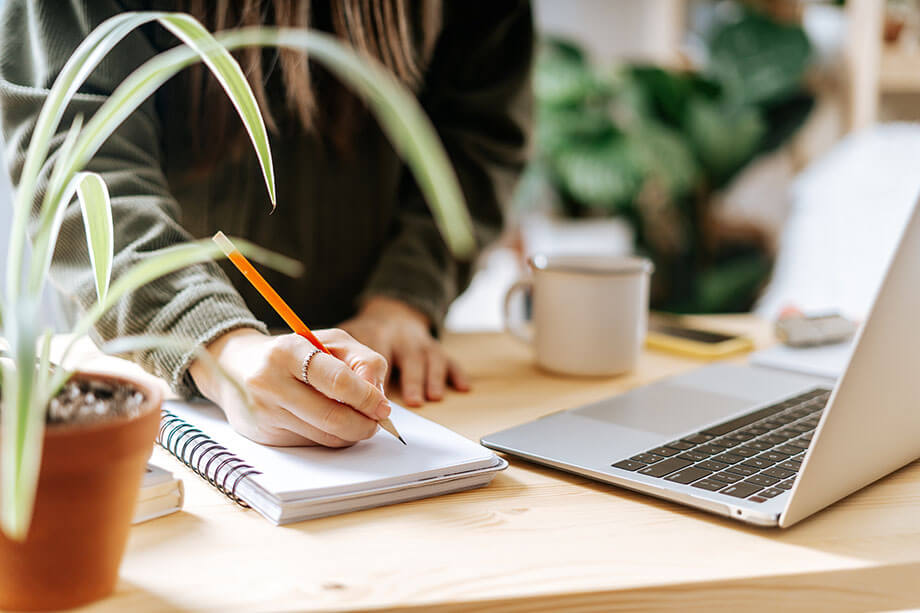 Series Saga Secrets A Writer's Guide to Success Woman writes in notebook in front of laptop while surrounded by desk plants