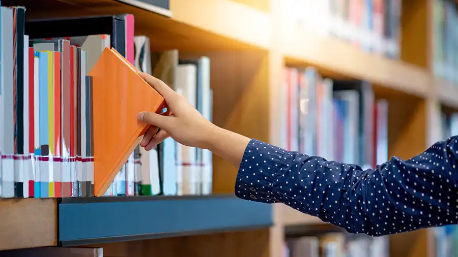 Genre Matters- How to Find the Best Fit for Your Book A woman's arm pulling an orange book from a library bookshelf.
