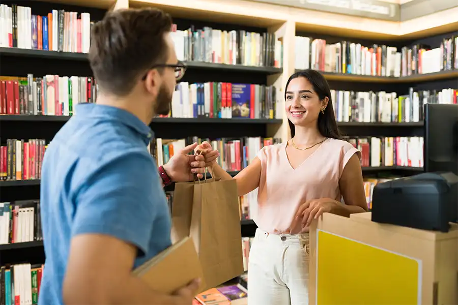 Ways You Are Hurting Your Book Sales A man in a blue polo purchasing books from a library. The woman librarian is handing over the books in a brown paper bag.